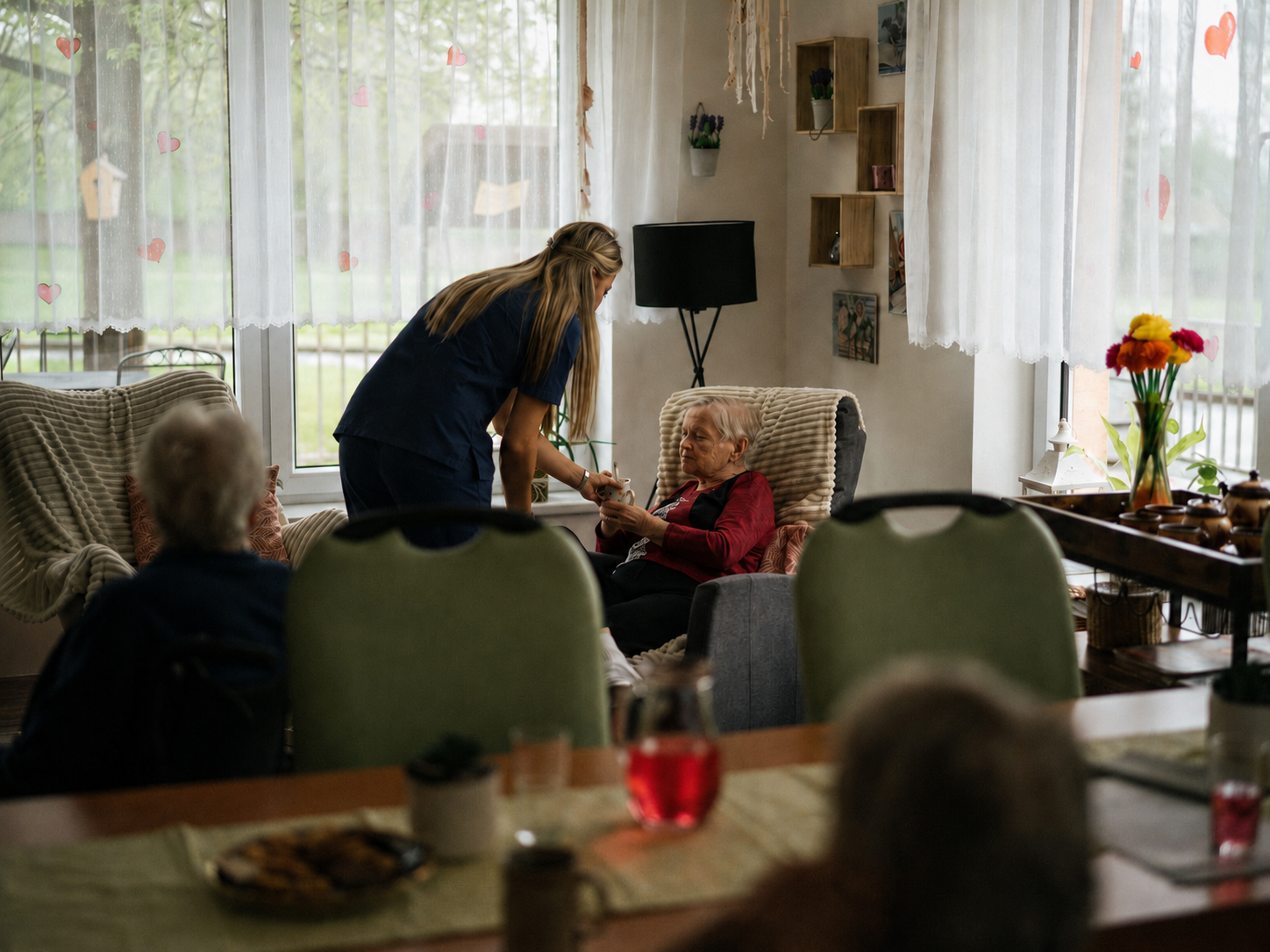 A care worker serves a hot drink to an elderly woman in a comfortable lounge, supporting calm companionship and consistent daily care.